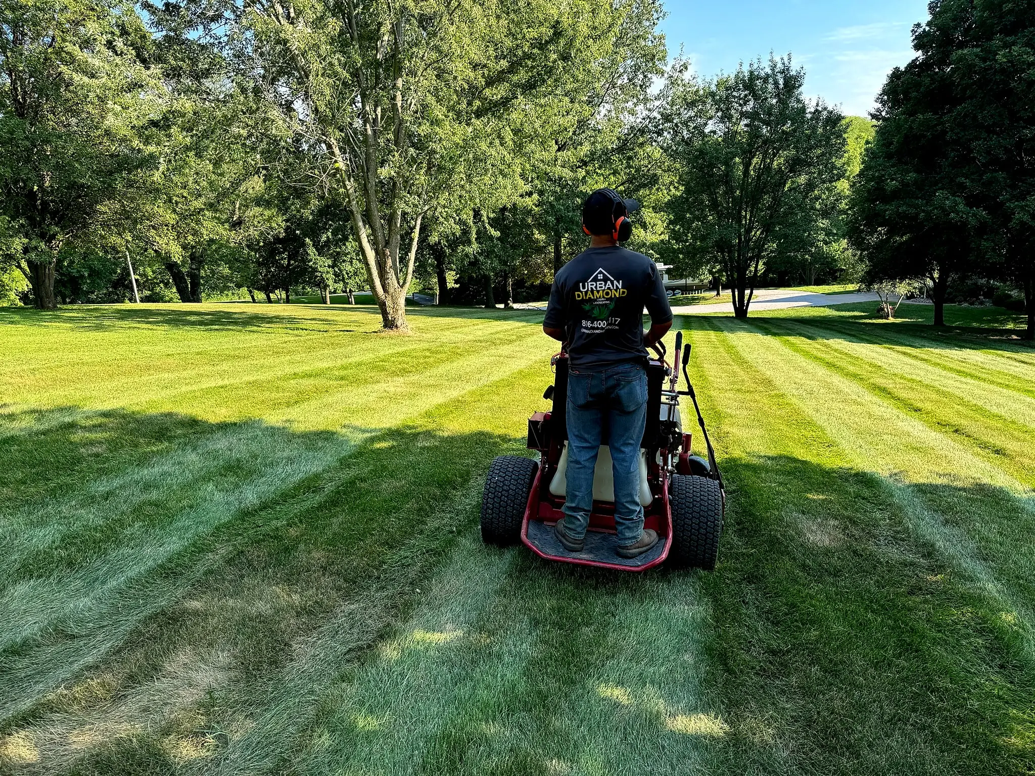 Urban Diamond crew member in a branded shirt mowing fresh lawn stripes on a stand-on mower